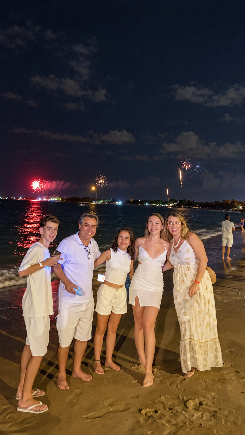 Família celebrando à beira-mar com reflexos dos fogos sobre a água