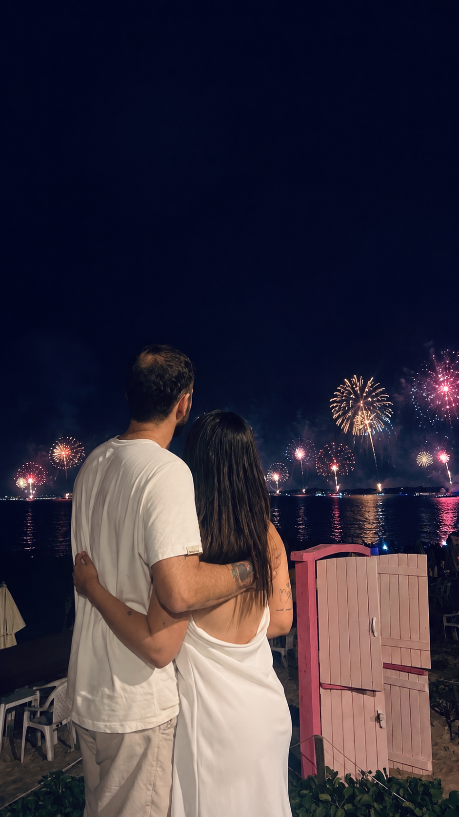 Casal abraçado diante do mar e dos fogos de artifício na virada do ano