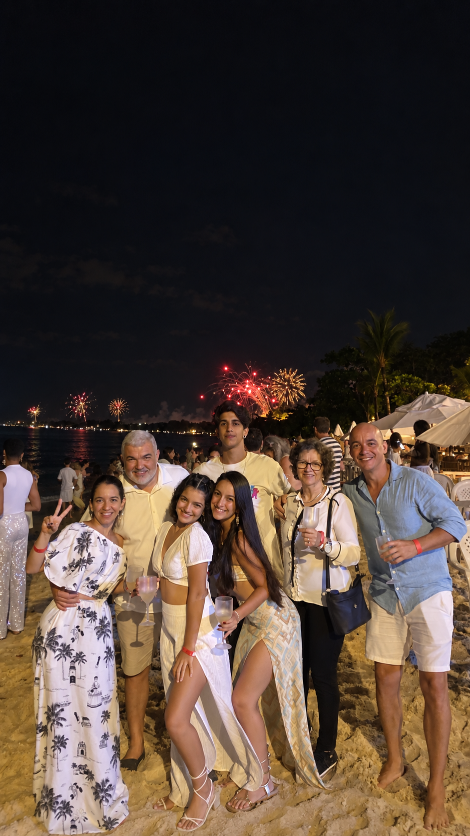 Grupo reunido na areia com fogos de artifício iluminando o litoral ao fundo